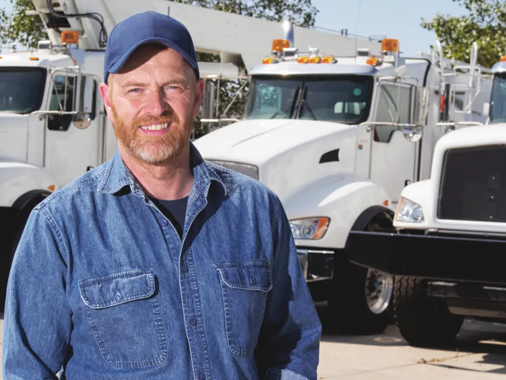 A smiling middle-aged man standing in front of a row of trucks in the outdoor sunshine.
